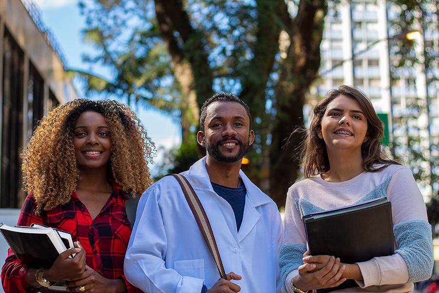 Three students in a row smiling, two students holding textbooks, 和 one student holding backpack strap.